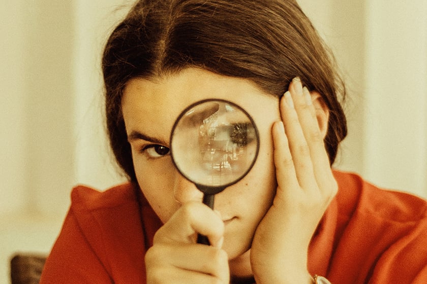 Woman looks through a magnifying glass, at the camera. Close crop on her face.