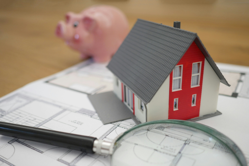 Model of a house placed on top of a housing blueprint with a piggy bank in the background
