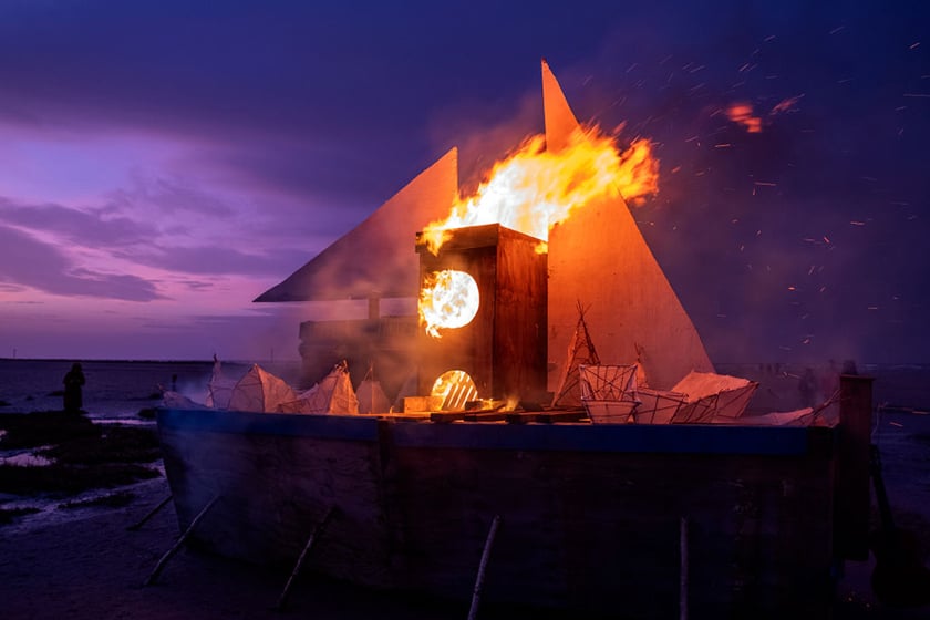 A bonfire in the shape of a boat, on a beach against a purple sky.