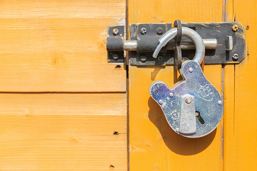 Close up shot of a strong but weatherbeaten padlock. It's open, and the bright yellow door it is supposed to be holding locked is slightly ajar. 